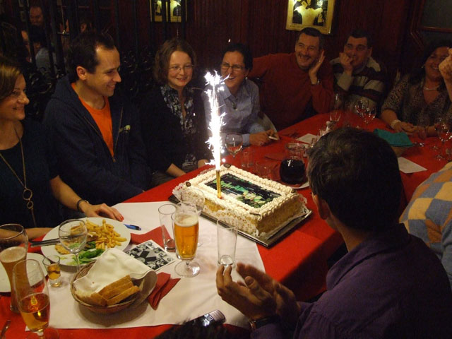 Az ünnepeltek egy asztal körül ülve nézik a szülinapi tortán égő tüzijáték-gyertyát.