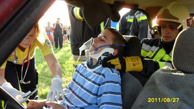 Doktornő lát el egy szimulált sérült gyermeket, aki egy autó ülésében ül. Mögötte egy tűzoltó segít az ellátásban.