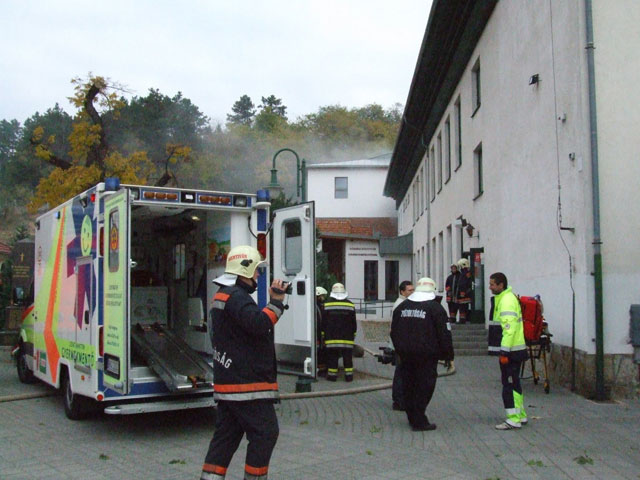 A Gyermekrohamkocsi nyitott hátsó ajtóval az iskola mellett áll. Körülötte tűzoltók és a személyzet egyik tagja.
